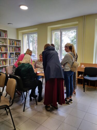 Renginys bibliotekoje. Žmonės stovi, sėdi prie stalo.