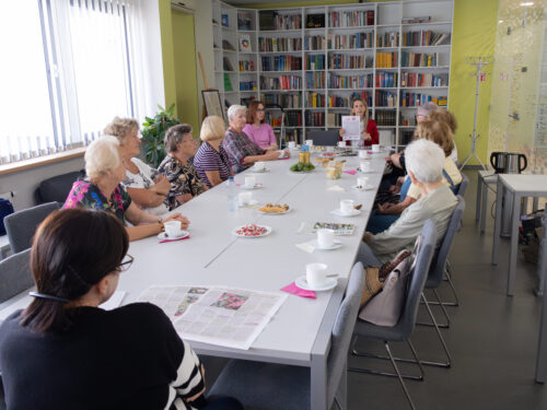 Penktadienio renginys "Spauda su kava". Prie stalo susėdę dvylika moterų, varto vietos spaudą, geria arbatą ar kavą iš baltų puodelių. Sausainiai, saldainiai, kava, arbata, spauda.