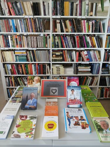 Bibliotekos erdvė. Nuotraukos gale lentyna pilna knygų. Priekyje stalas, spaudinių paroda „Stiprinkime sveikatą“. Išdėliotos knygos.